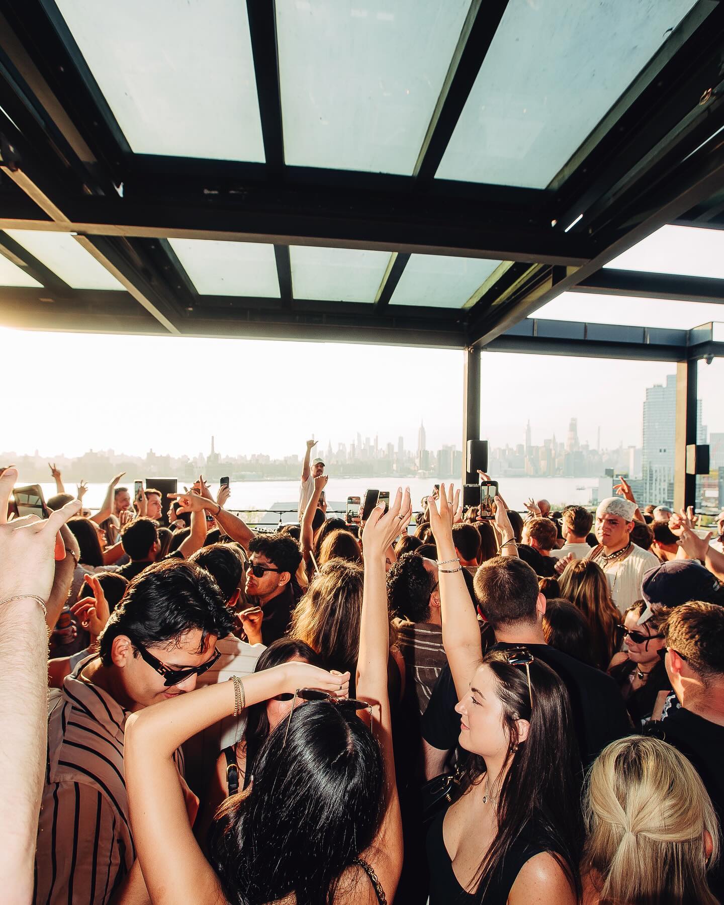 A crowded group of people are dancing and raising their hands at an outdoor rooftop party, with a city skyline visible in the background.