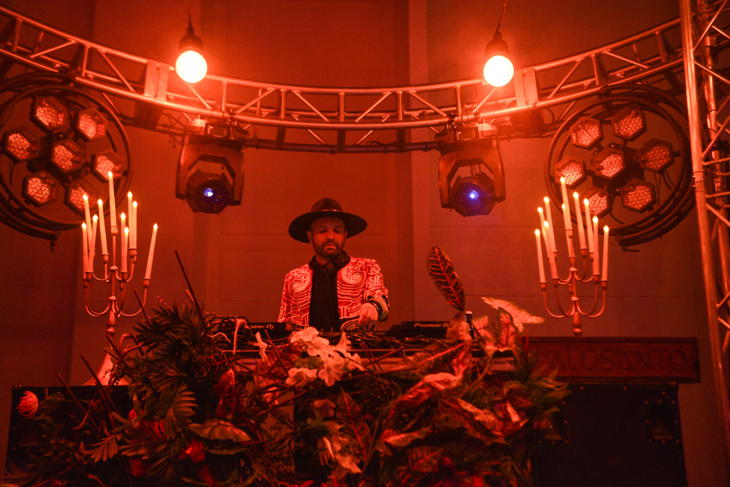 A DJ wearing a hat and patterned jacket performs on stage surrounded by red lighting, candelabras, and floral decorations.