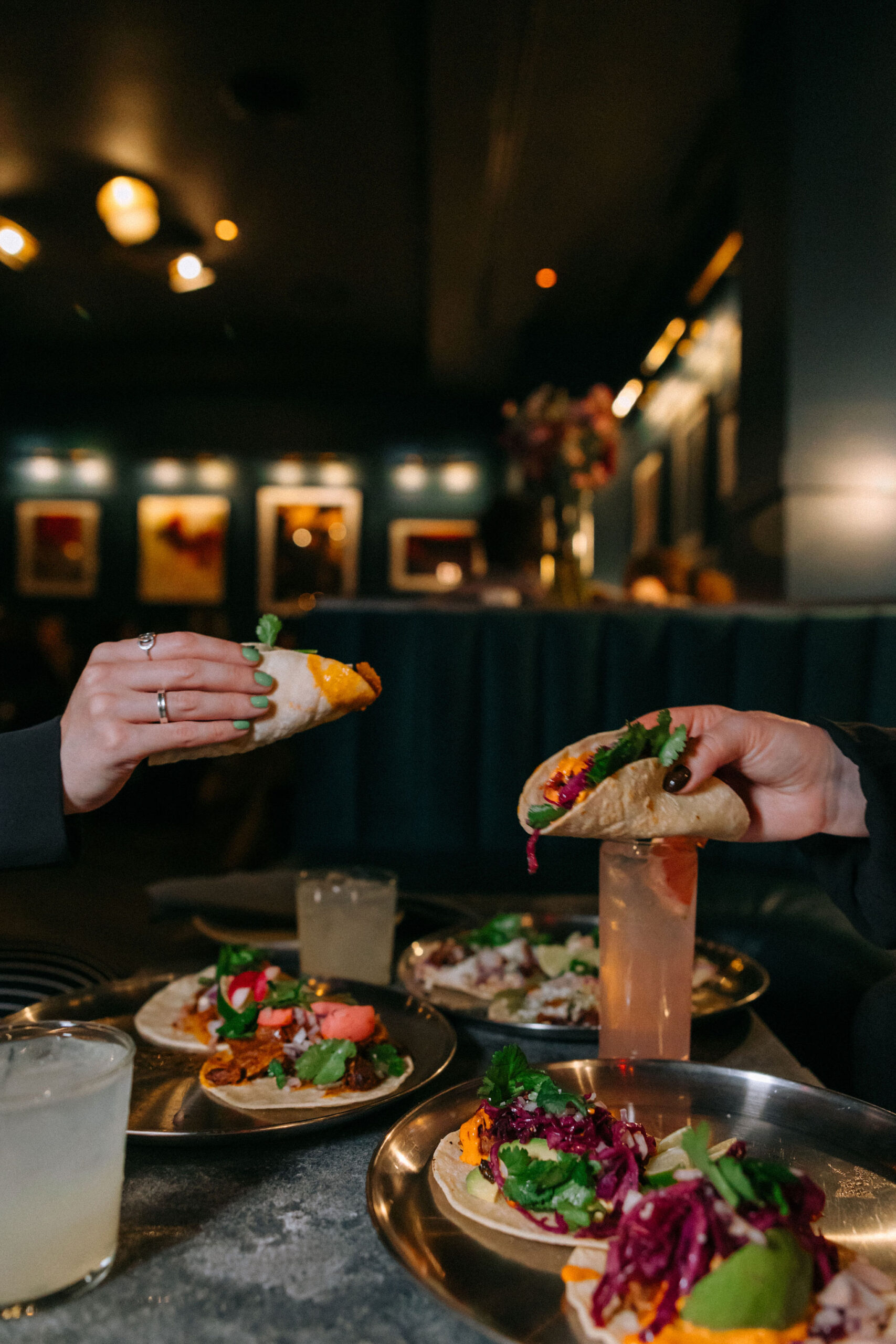 Two hands holding tacos over a table with multiple plates of tacos and drinks in a dimly lit restaurant. The background shows a stylish, dark interior with framed pictures on the walls.