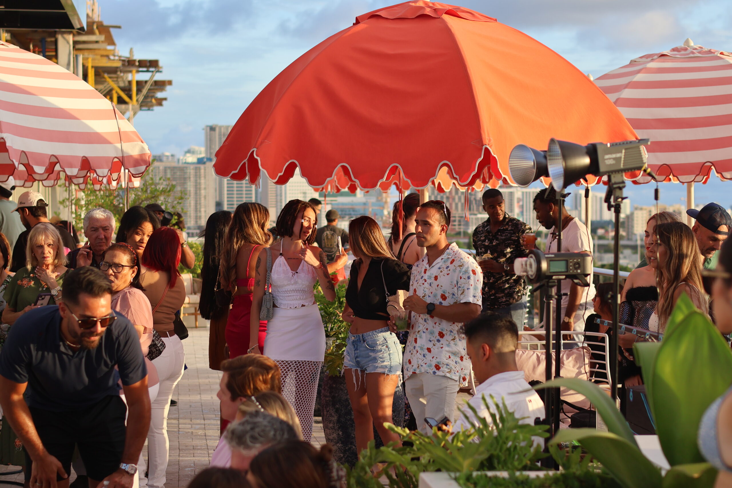 A crowded rooftop gathering under orange umbrellas, with people socializing, chatting, and enjoying the view of a cityscape in the background.