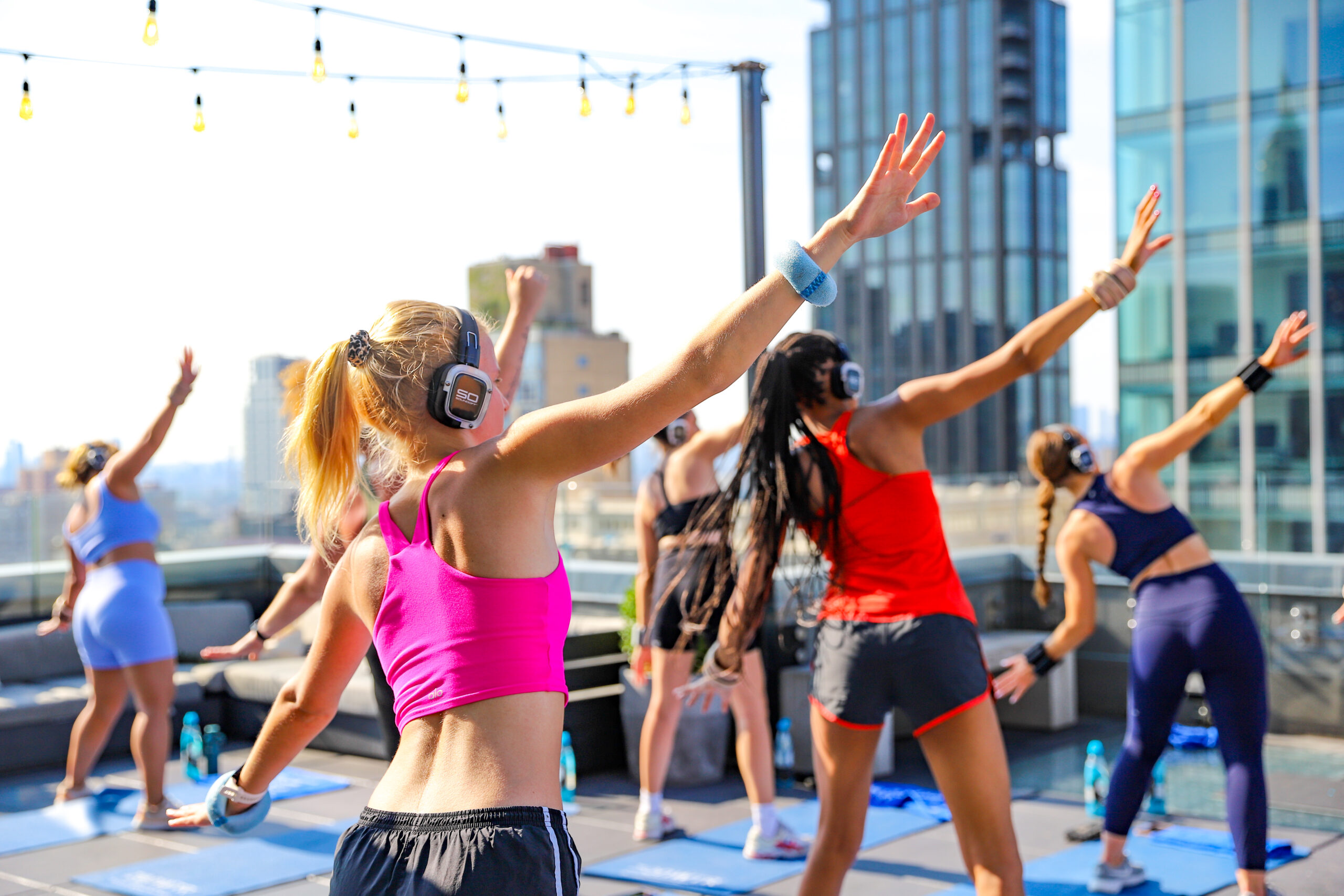 A group of people participate in a rooftop exercise class, wearing wireless headphones and workout attire, with city buildings in the background.