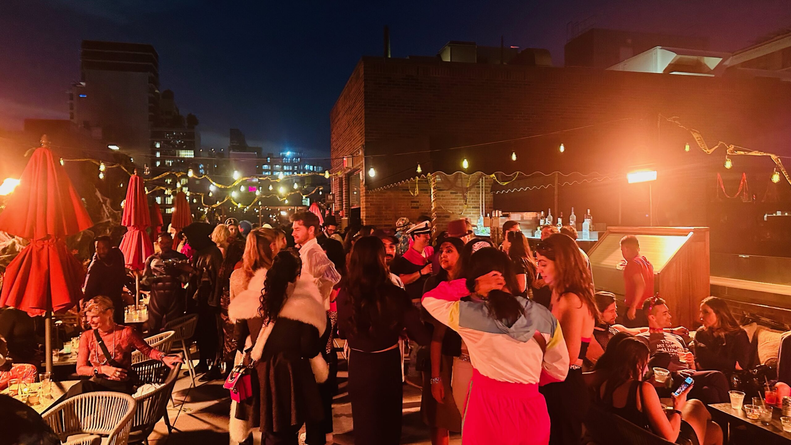 A nighttime rooftop party with people socializing, colorful string lights, and patio furniture in a city setting.