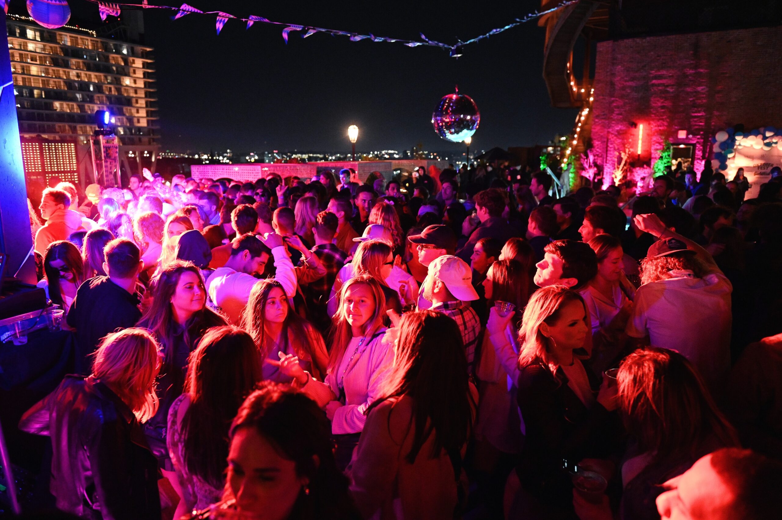 A large crowd of people gather for a night party on a rooftop, illuminated by vibrant colorful lights.