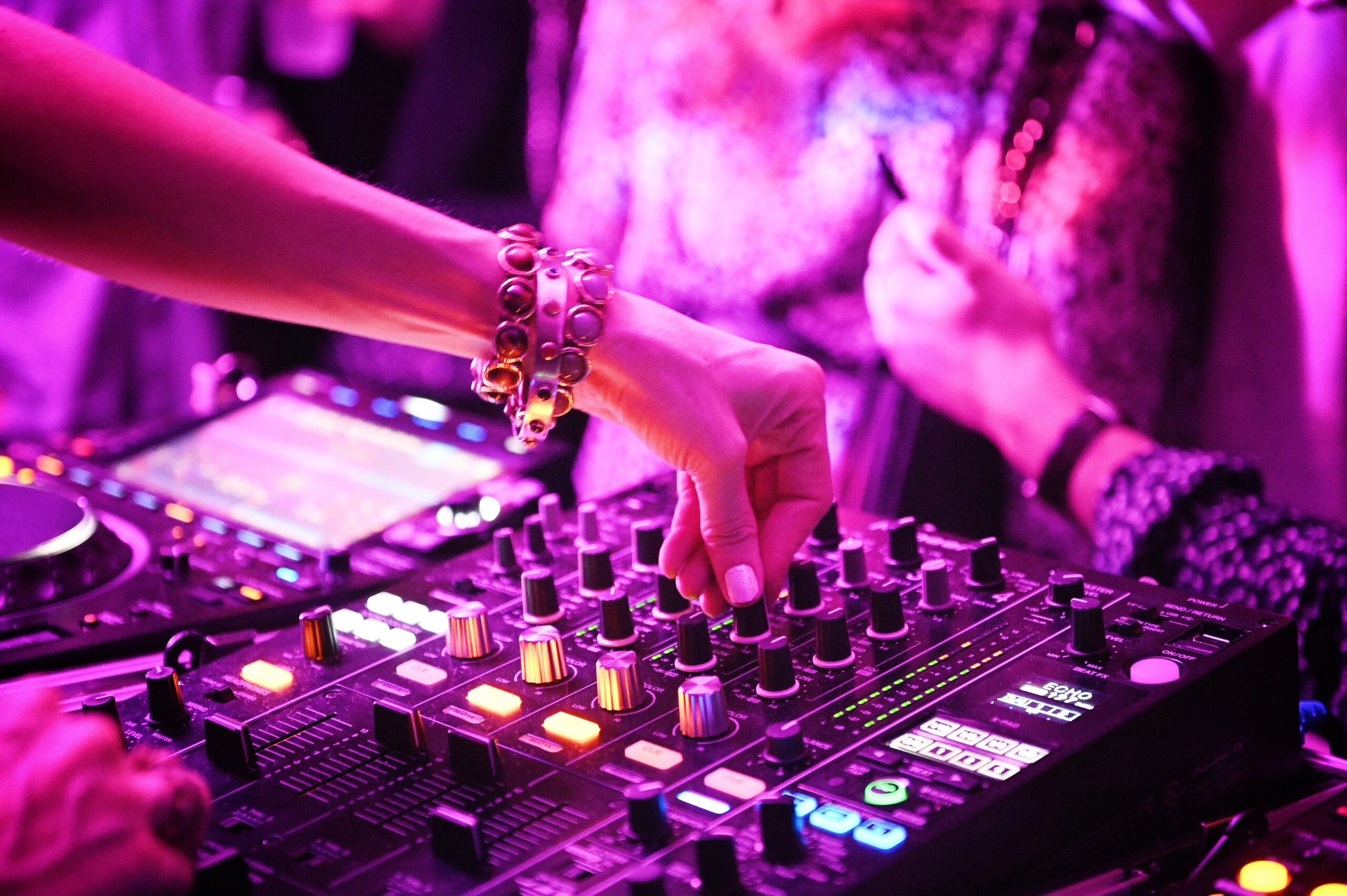 A DJ's hand adjusts knobs on a mixing console at a vibrant, purple-lit event.