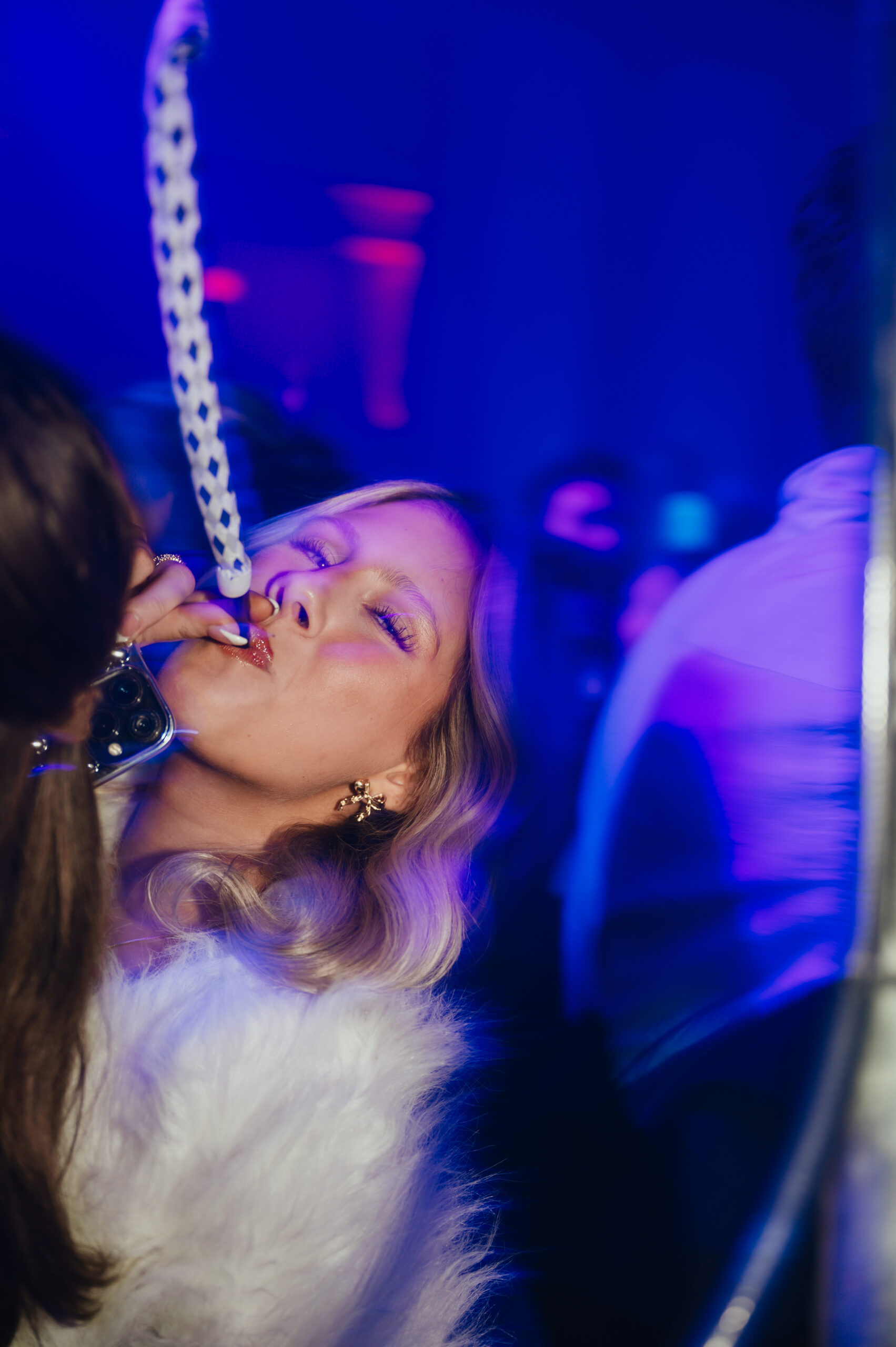 Person in a fur coat holds a patterned straw to their mouth in a dimly lit, blue-hued nightclub setting.