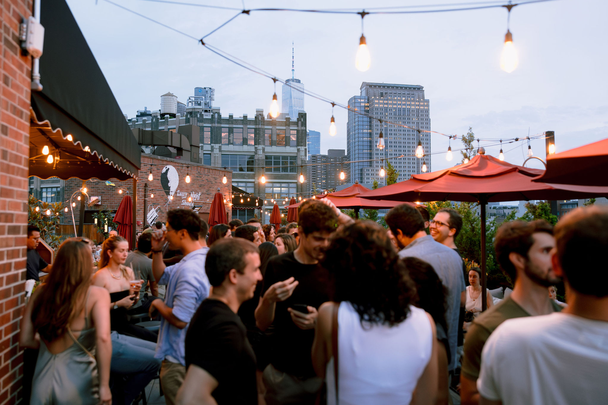 A group of people socializing on a rooftop with string lights, red umbrellas, and city buildings in the background.