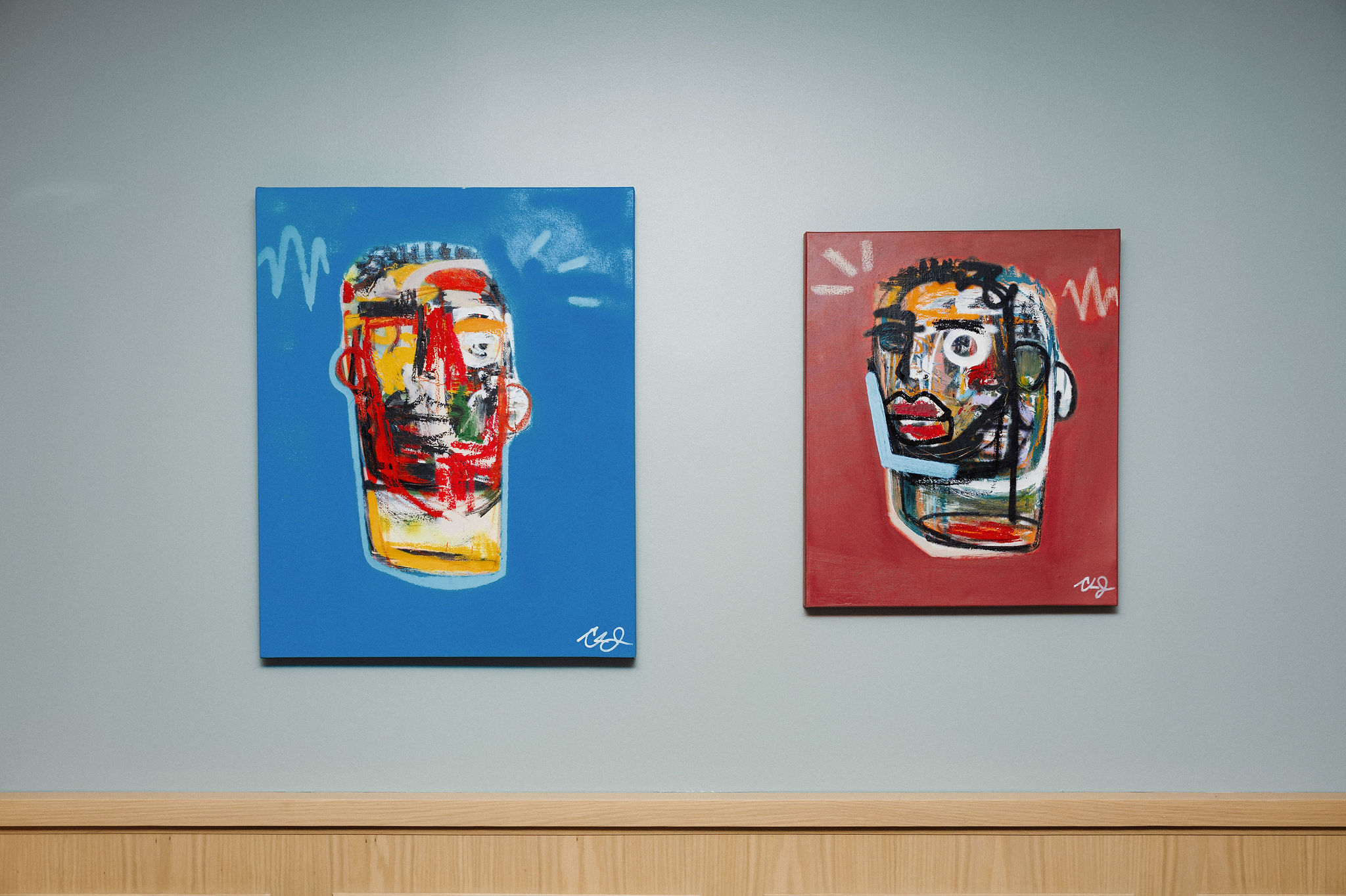 Two abstract paintings featuring stylized human faces with vivid, bold colors; one on a blue background, the other on red, hung on a neutral wall with a wooden trim below.