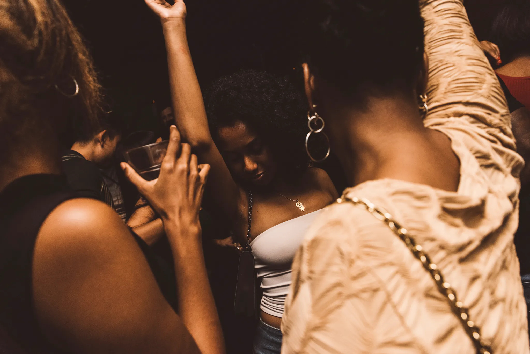 People dancing closely in a dimly lit space. One person holds a drink, another raises their arm. They are wearing casual outfits, with some jewelry visible.